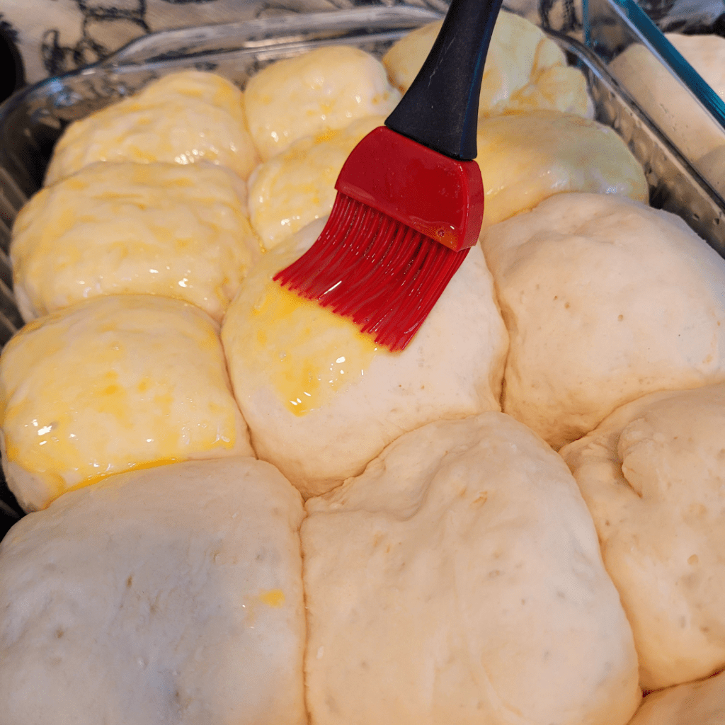 Image illustrates proofed dinner rolls being brushed with an egg wash.