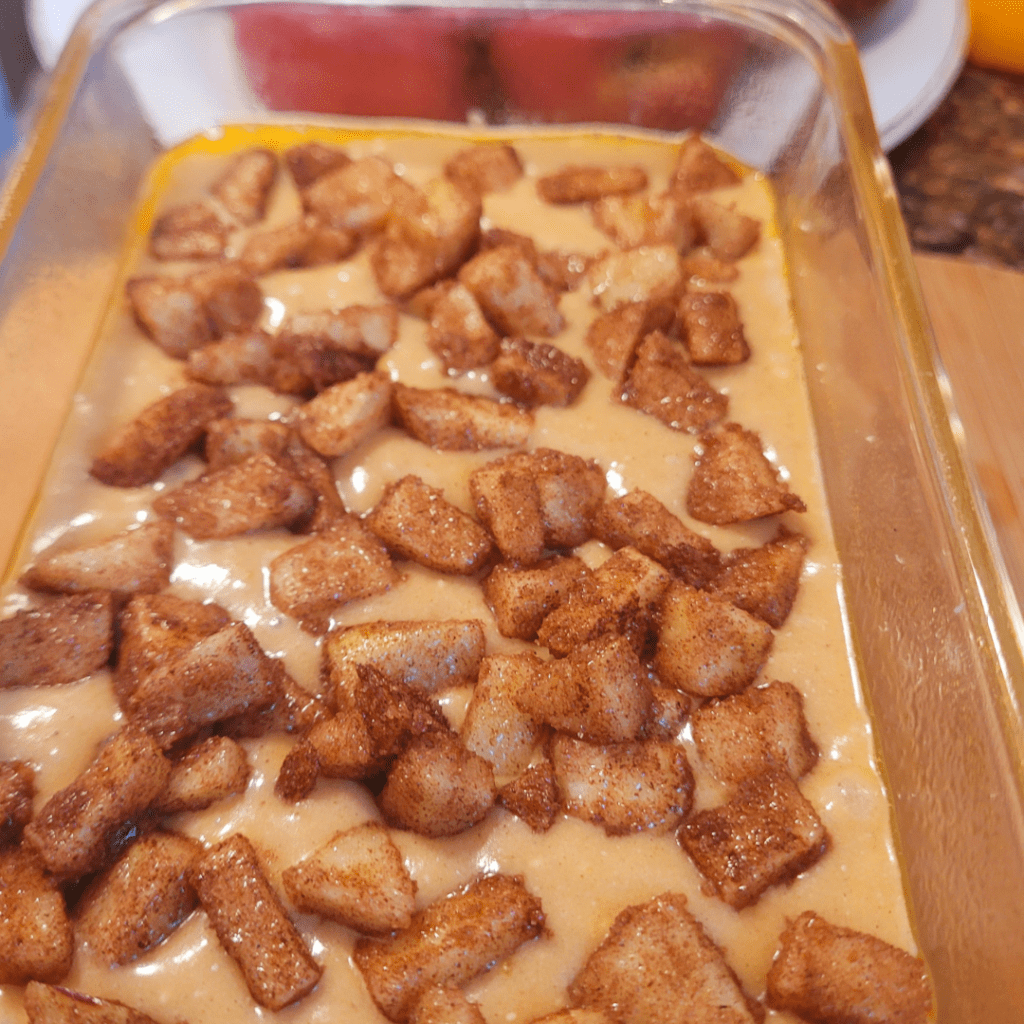 Image illustrates diced apples covered in cinnamon and sugar on top of bread batter for an apple fritter bread recipe. 