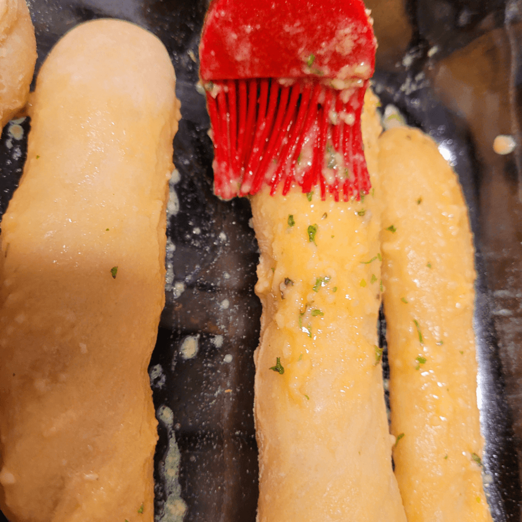 Image illustrates breadsticks being brushed with a topping for breadsticks.